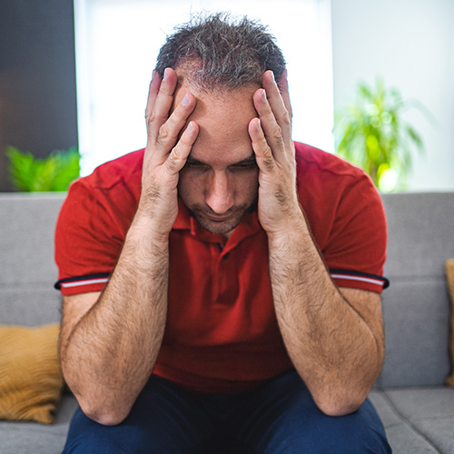 Hair Loss From Stress. A man sits on a couch holding his hands to his head in stress.